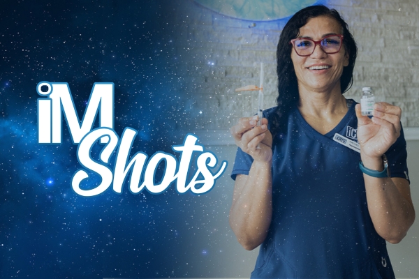 A smiling woman wearing blue scrubs and red-framed glasses holds two small lab vials